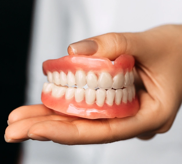 Hand holding a complete denture model displaying full upper and lower artificial teeth.