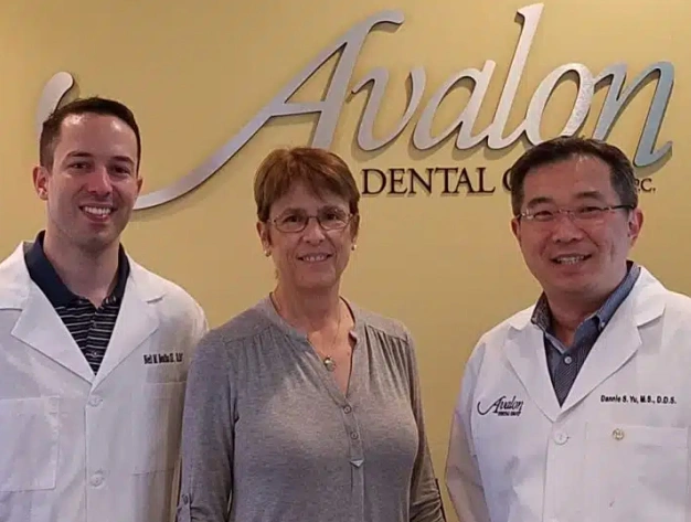 Three dental professionals posing together in front of Avalon Dental Group logo at their office.