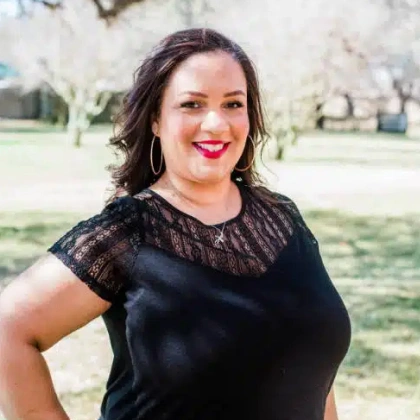 Woman smiling outdoors wearing black lace top with hoop earrings, park setting background.