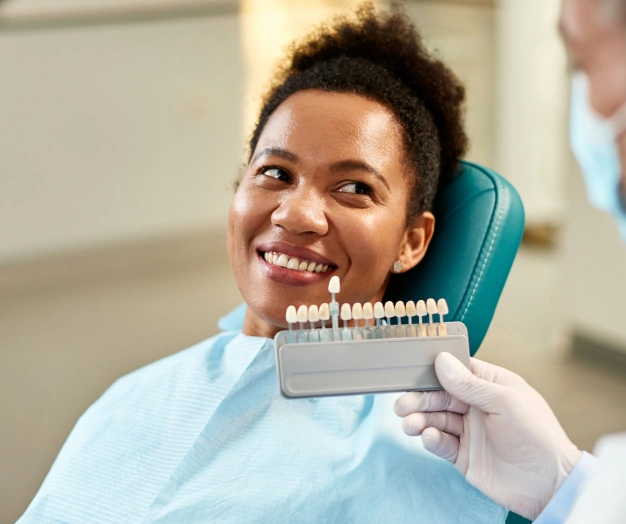 Young man in dental chair smiling while holding tooth shade guide during dental consultation.