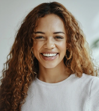 Smiling woman with curly brown hair wearing light ribbed sweater in bright indoor setting.