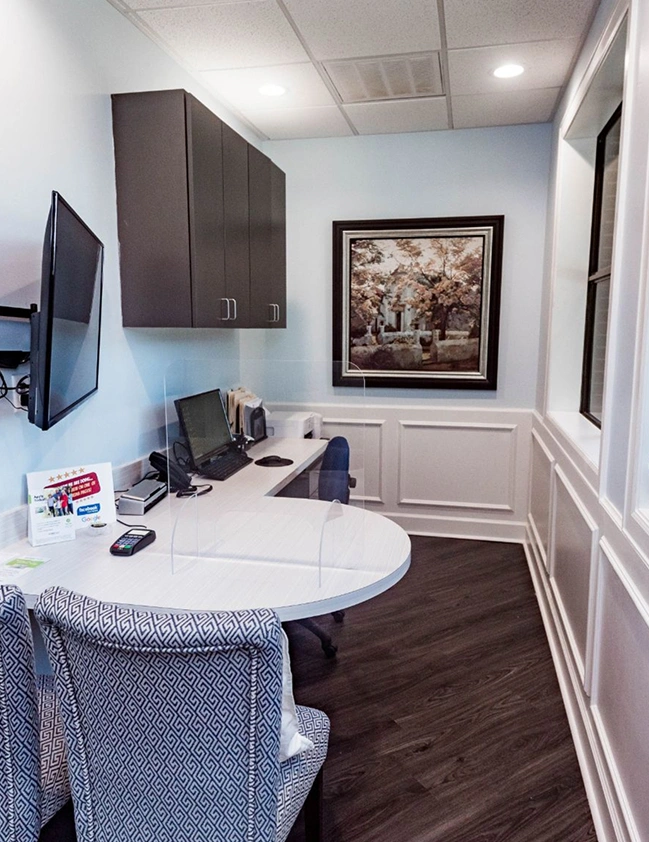 Modern dental office reception area with curved white desk, computer equipment, and framed artwork on walls.