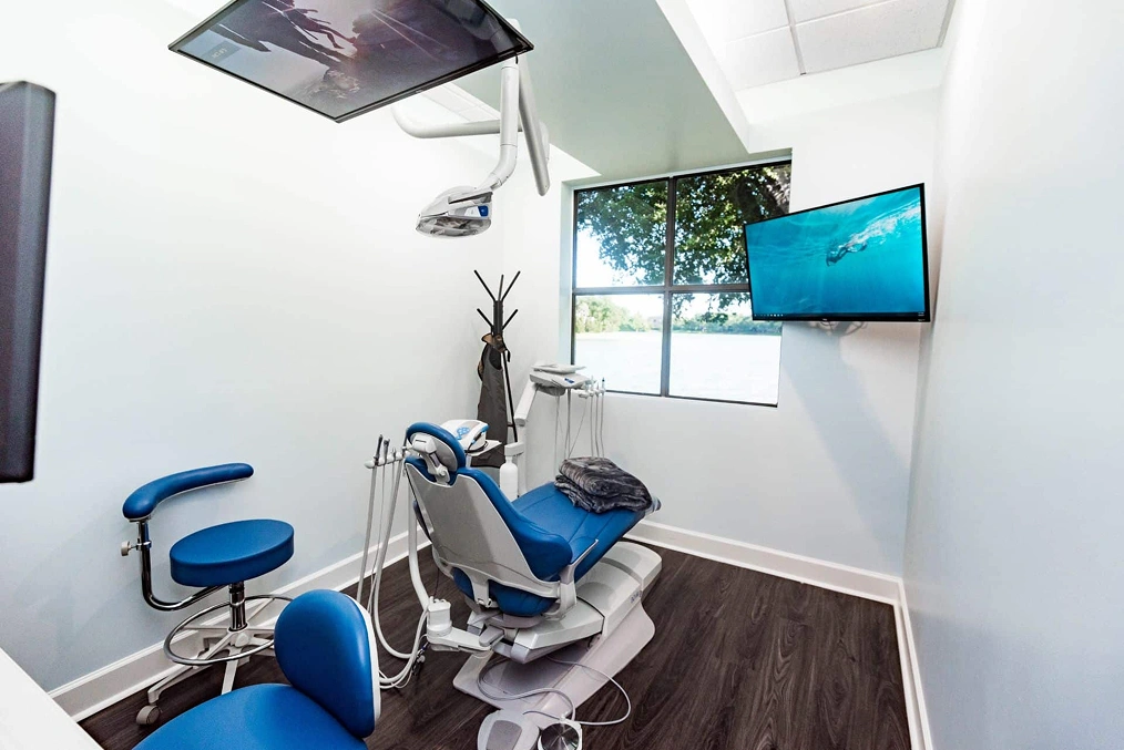 Modern dental operatory with blue patient chair, equipment, and mounted monitor in bright clinical space.