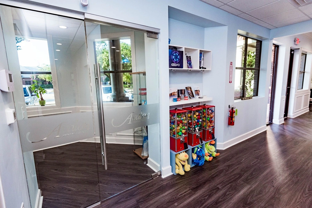 Modern dental office reception area with glass doors, white walls, and dark flooring.