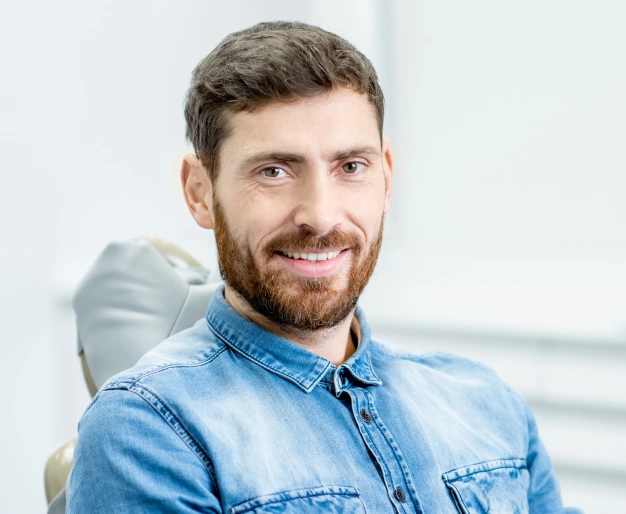 Professional man in blue denim shirt smiling at camera in modern dental office setting.