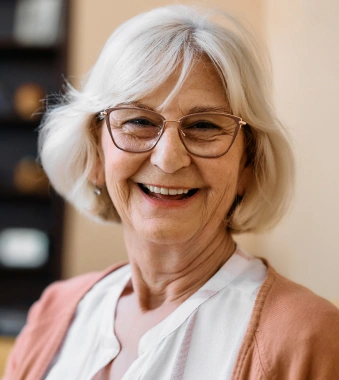 Smiling older woman with white hair and glasses wearing pink cardigan in dental office setting.