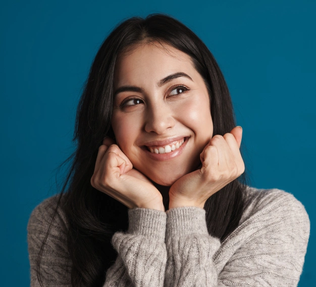Young woman smiling at camera with hands near face against blue background, dental practice portrait.