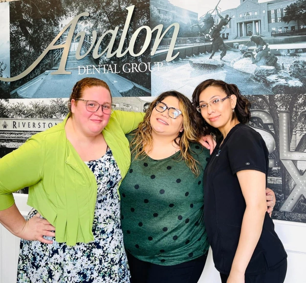 Three women posing together at Avalon Dental Group office with branded backdrop behind them.