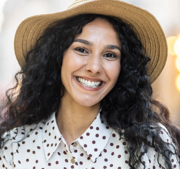 Young woman smiling at camera wearing straw hat and polka dot shirt, indoor setting.