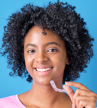 Smiling woman holding clear aligner against blue background for dental care.