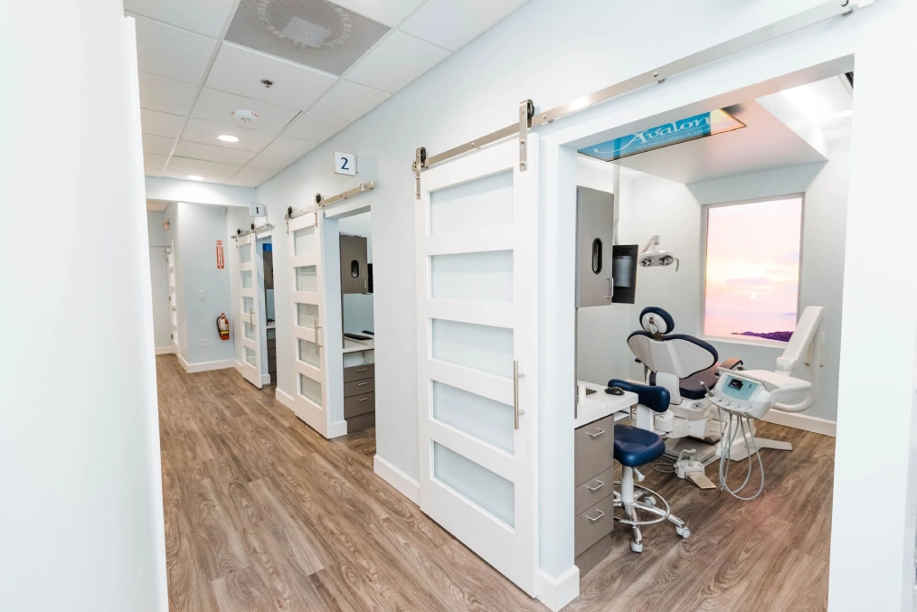 Modern dental office interior with treatment rooms, white walls, wood flooring, and clinical equipment visible.