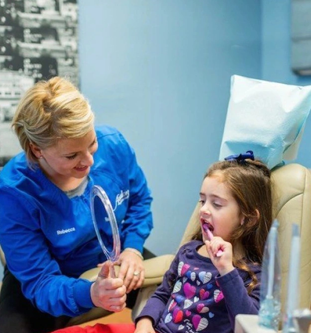 Female dental hygienist in blue scrubs examines young girl's teeth in clinical dental office setting.