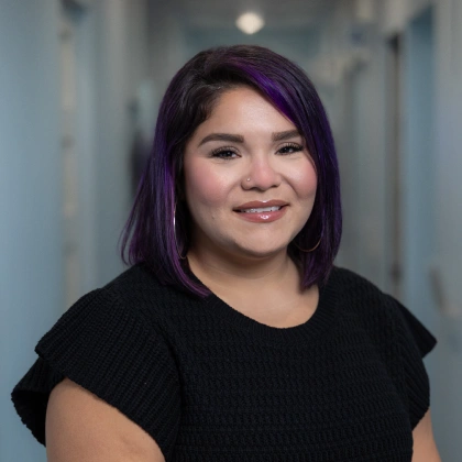 Young woman with purple hair smiling at camera in clinical hallway setting.