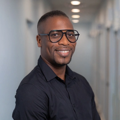 Professional man wearing glasses and dark shirt smiling in modern dental office hallway.