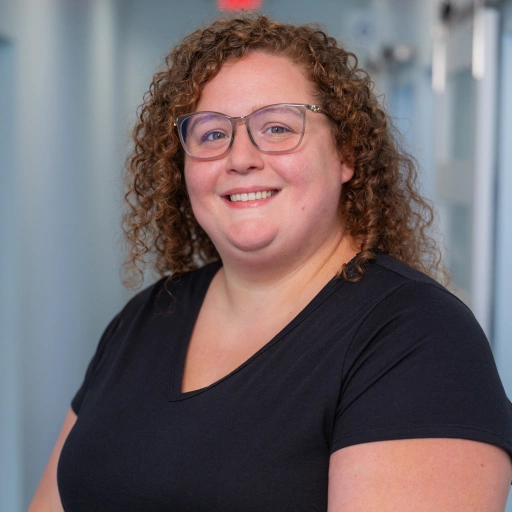Woman wearing glasses and black shirt smiles at camera in dental office hallway.