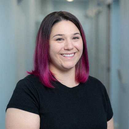 Young woman with magenta-streaked dark hair smiling in modern dental office hallway.