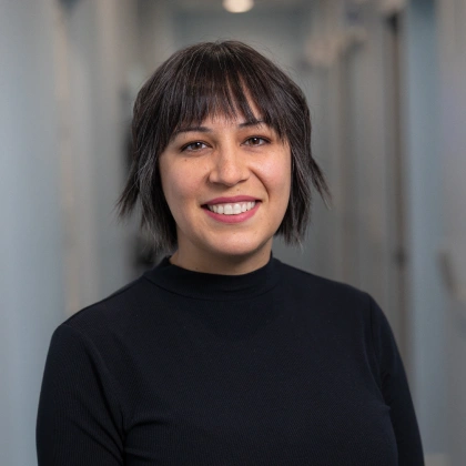 Professional headshot of smiling woman in black turtleneck against blurred office hallway background.