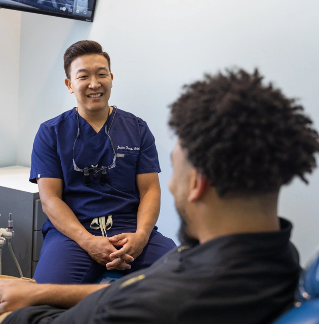 Dentist in blue scrubs smiling while consulting with patient in dental office.