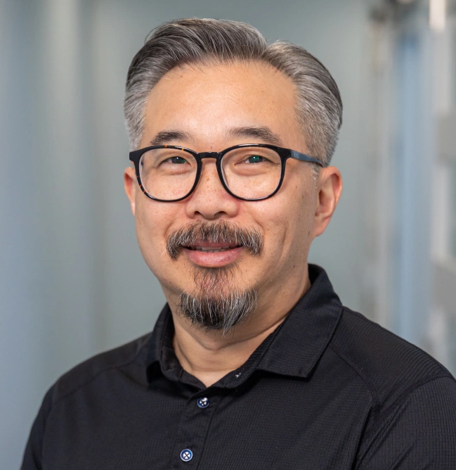 Professional headshot of man wearing glasses and black polo shirt against blurred office background.