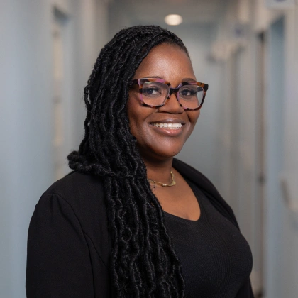 Professional headshot of smiling woman with glasses in modern dental office hallway.