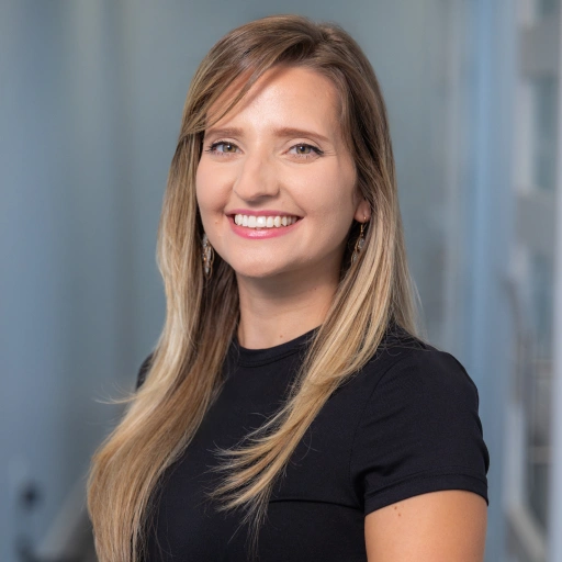 Professional headshot of smiling woman in black shirt against blurred office background.