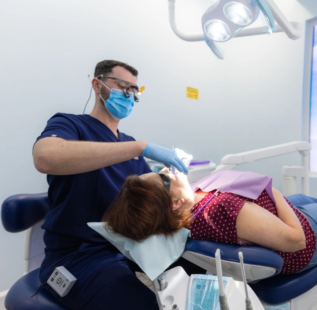 Dentist in blue mask and gloves examines patient in dental chair under overhead light.