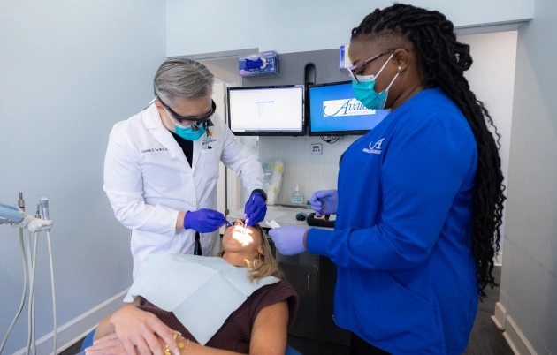 Dentist in white coat and mask examines patient's teeth with assistant in blue scrubs.