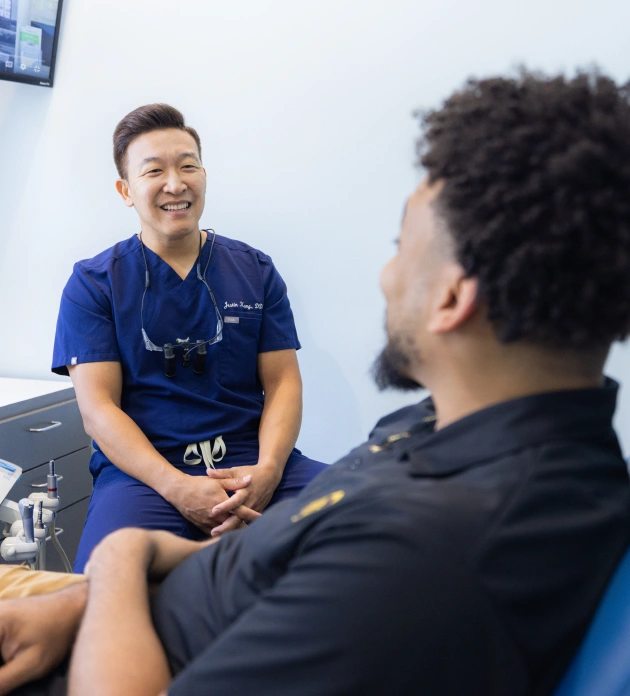 Dental professional in blue scrubs smiling while consulting with male patient in clinical setting.