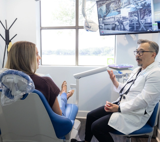 Male dentist consulting with female patient in bright dental office with large windows and artwork.