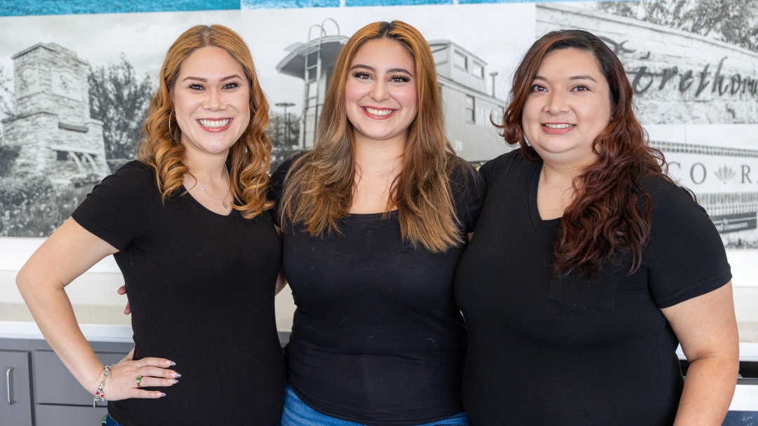 Three female dental professionals smiling at camera in modern office setting with blue background.