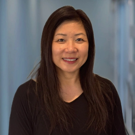 Professional headshot of smiling woman with long dark hair wearing dark top against blue background.