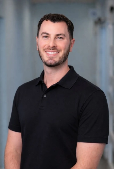 Professional headshot of smiling man in black polo shirt against blurred blue background.