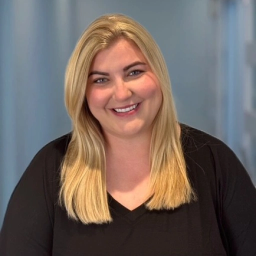 Professional headshot of smiling woman with blonde hair in black top against blue background.