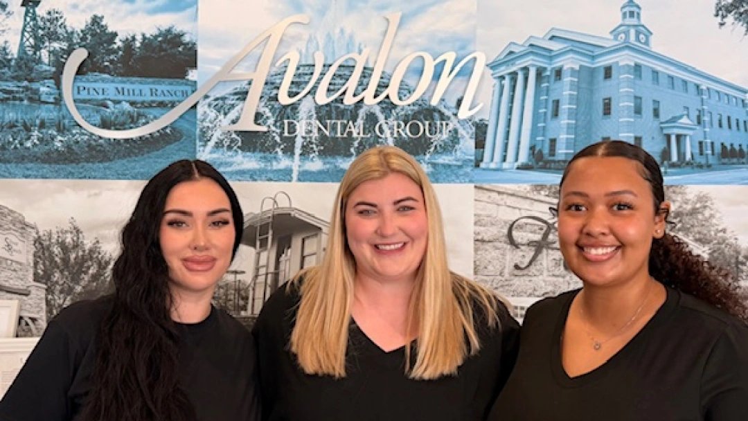 Three dental professionals smile at camera with Avalon Dental Group branding and office building backdrop